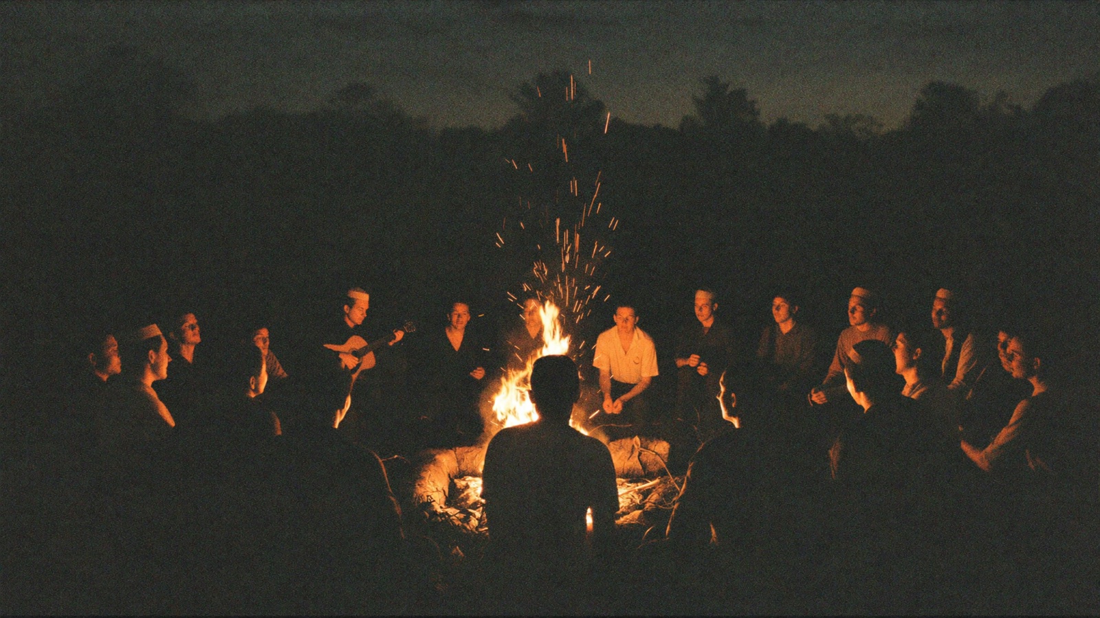 A wide circle of people seated on logs around a large campfire at night, sparks rising into a black sky